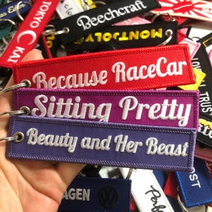 May include: Three colorful keychains with embroidered text. The top keychain is red with white text that reads "Because RaceCar". The middle keychain is purple with white text that reads "Sitting Pretty". The bottom keychain is purple with white text that reads "Beauty and Her Beast".