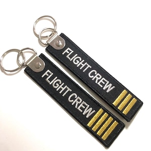 Op de afbeelding: Twee zwarte sleutelhangers met zilveren metalen sleutelringen. De sleutelhangers zijn gemaakt van zwart stof met witte geborduurde tekst die "FLIGHT CREW" luidt. De sleutelhangers hebben drie gouden strepen.