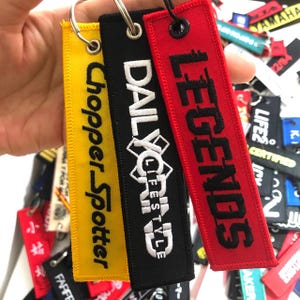 May include: Three black, red, and yellow fabric keychains with embroidered text. The black keychain says "Daily X Life", the red keychain says "Legends", and the yellow keychain says "Chopper Spotter".