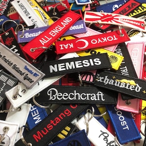 May include: A collection of colorful keychains with embroidered text. The keychains feature various designs and phrases, including "All England", "Nemesis", "Tokyo", "Exhausted", and "Beechcraft".