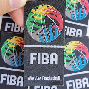 Könnte beinhalten: Ein schwarzer und weißer Patch mit dem FIBA-Logo. Das Logo ist ein Basketball, der von Händen umgeben ist, die die Welt repräsentieren. Der Text "FIBA" ist in weißen Buchstaben und der Text "We Are Basketball" ist in weißen Buchstaben unter dem Logo.