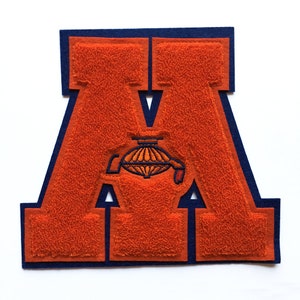 May include: A large orange felt letter M with a navy blue outline. The letter is a varsity style patch with a small embroidered image of a ship's wheel inside the letter.
