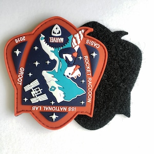 Könnte beinhalten: Ein roter, weißer und blauer gestickter Aufnäher mit dem Text "ISS NATIONAL LAB", "GROOT", "ROCKET RACCOON", "CASIS", "MARVEL" und "2016". Der Aufnäher zeigt ein stilisiertes Bild einer Raumstation und einer Figur aus dem Marvel Cinematic Universe.