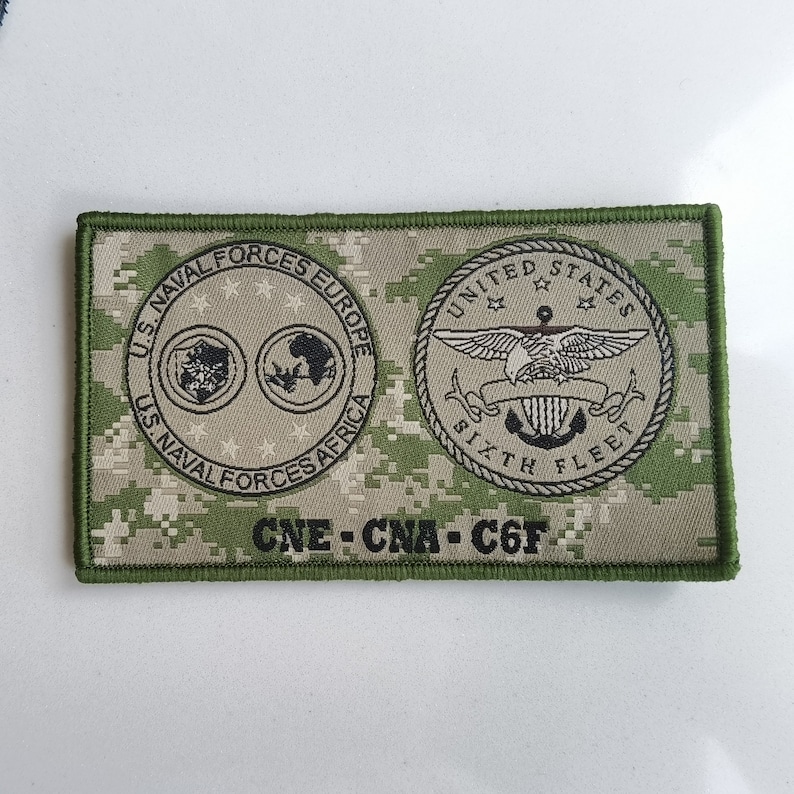 May include: A green and tan camouflage patch with the text "U.S. NAVAL FORCES EUROPE", "US. NAVAL FORCES AFRICA", "UNITED STATES", "SIXTH FLEET", and "CNE-CNA-C6F".