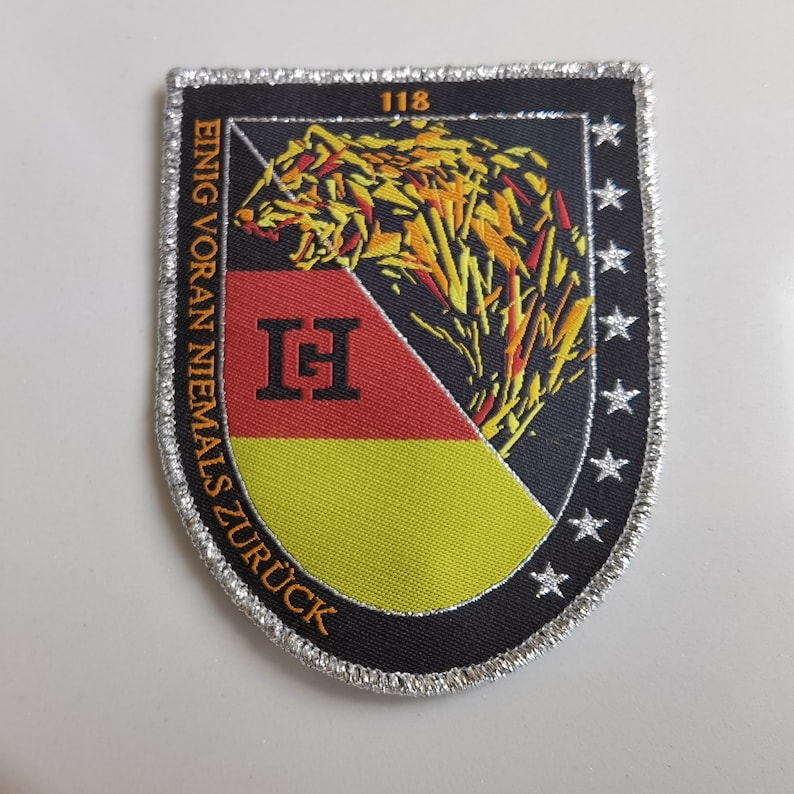 May include: A black and silver embroidered patch with a wolf's head, a German flag, and the text "EINIG VORAN NIEMALS ZUR&Uuml;CK" and "118" in orange. The patch has a silver border with stars.