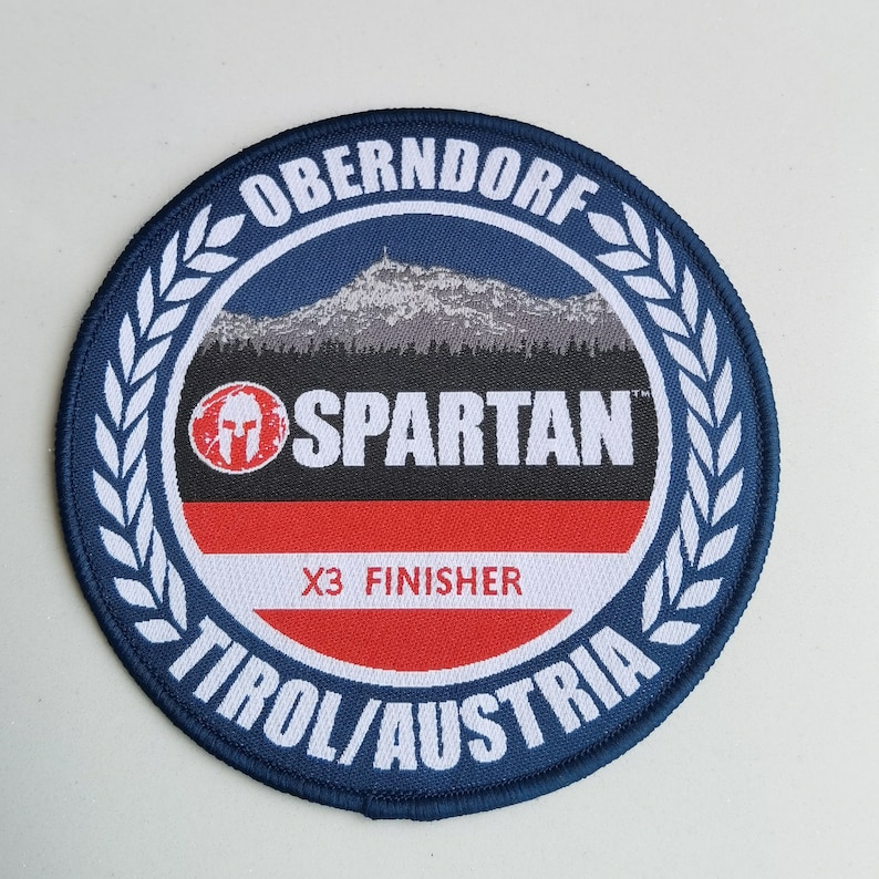 May include: A blue and white circular patch with the text "OBERNDORF", "SPARTAN", "X3 FINISHER", and "TIROL/AUSTRIA". The patch features a mountain range and a Spartan helmet logo.