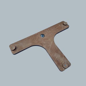 May include: A T-shaped wooden component featuring a smooth, light brown finish with visible wood grain. The piece includes three small, circular wooden spacers attached at each end and a central metal fastener, creating a flat, structural element.