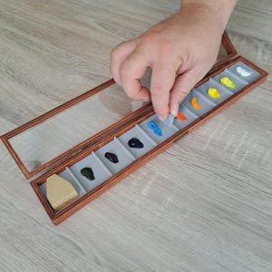 Könnte beinhalten: Eine hölzerne Künstlerpalette mit einem Deckel aus klarem Glas, die verschiedene Farben enthält. Die Box ist geöffnet, und eine Hand greift hinein, um die Farbe anzupassen. Die Farben umfassen Blau, Schwarz, Grün, Gelb, Orange und Weiß.