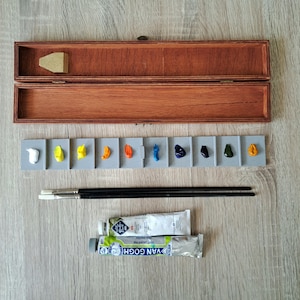 Op de afbeelding: Een houten kunstschildersdoos met een scharnierend deksel, met een grijs palet met klodders van verschillende kleuren, twee penselen en twee tubes Van Gogh-verf. Het palet bevat witte, gele, oranje, blauwe en groene verf.