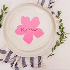May include: A white ceramic plate with a smaller plate on top, featuring a pink flower design with the name "Patricia" in cursive. Green sprigs and a blue and white striped cloth add a decorative touch.