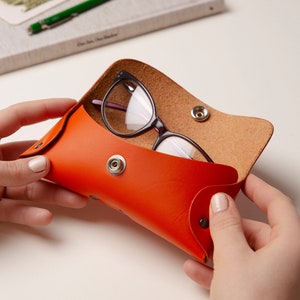 May include: Orange leather eyeglasses case with a snap closure. The case is open and a pair of eyeglasses is inside.