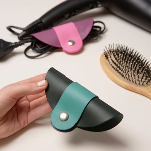 May include: A set of three leather cord organizers in green, pink, and purple. The organizers are shaped like half-circles and have a button closure.