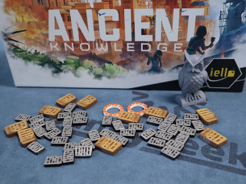 May include: A game board box with the text "ANCIENT KNOWLEDGE" and an owl figurine. The box is on a blue surface with a variety of small wooden and gray tiles scattered around it. The tiles are rectangular and have a grid pattern.