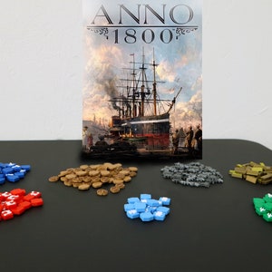 Puede incluir: Un tablero de juego con varias piezas de juego de colores, incluyendo azul, rojo, azul claro, verde, marrón y gris. La caja del juego está en el fondo y dice "Anno 1800".