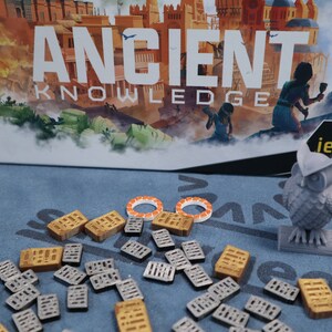 May include: A board game called "Ancient Knowledge" with a gray owl figurine, orange and white tokens, and gray and gold building pieces.