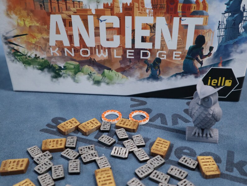 May include: A board game called "Ancient Knowledge" with a gray owl figurine, orange and white tokens, and gray and gold building pieces.