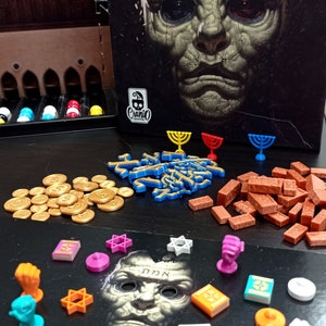 Puede incluir: Un tablero de juego con varias piezas de juego, incluyendo monedas de oro, ladrillos rojos, letras hebreas azules y pequeñas figuras. El tablero de juego presenta una ilustración en blanco y negro de la cara de un hombre con la palabra hebrea "אמת" (verdad) escrita en su frente.