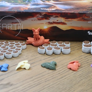 Può includere: Un gioco da tavolo chiamato "Earth" di InsideUp Games. Il gioco include una varietà di gettoni colorati, tra cui una piccola statuetta di volpe arancione, e una scatola con una scena di tramonto.