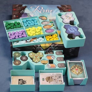 May include: A teal blue board game organiser with multiple compartments for storing game pieces and cards. The organiser is labelled "Rovic" and features a variety of compartments for storing different sized game pieces.