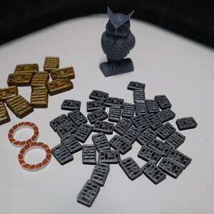 May include: A gray 3D printed owl figurine, a pile of gold-colored tiles, and a pile of gray tiles. The tiles are rectangular and have a textured surface.