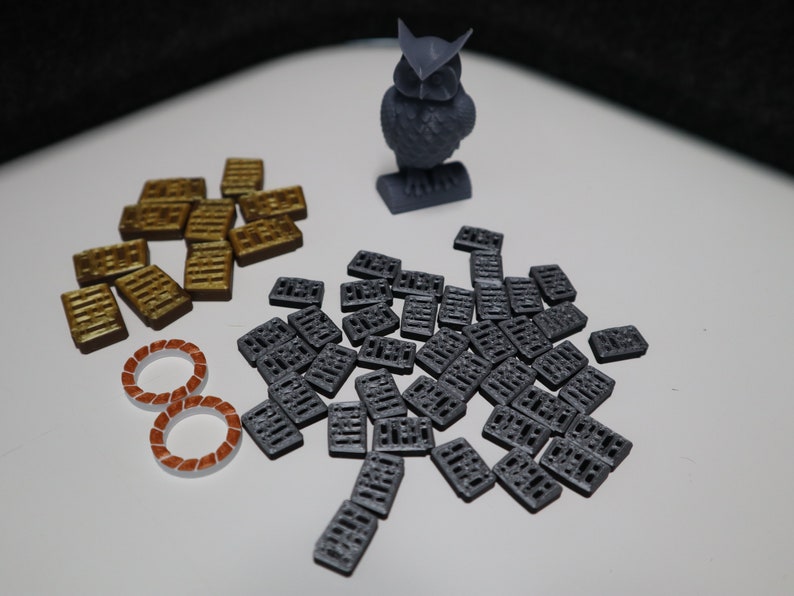 May include: A gray 3D printed owl figurine, a pile of gold-colored tiles, and a pile of gray tiles. The tiles are rectangular and have a textured surface.