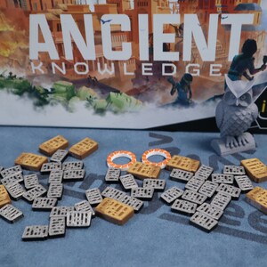 May include: A game board with the title "Ancient Knowledge" and a gray owl figurine. The game pieces are small, rectangular tiles with a grid pattern. Some tiles are brown and some are gray.