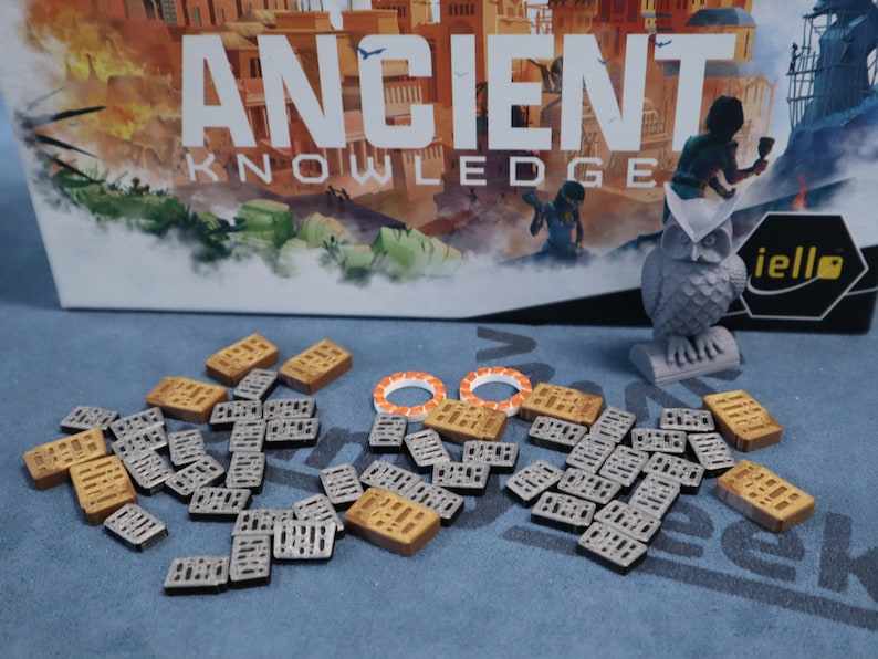 May include: A game board with the title "Ancient Knowledge" and a gray owl figurine. The game pieces are small, rectangular tiles with a grid pattern. Some tiles are brown and some are gray.