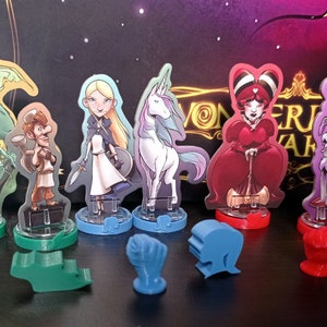 Puede incluir: Un conjunto de piezas de juego coloridas que incluyen un dragón verde, una niña de cabello castaño con una espada, un unicornio blanco, una reina roja con una corona, un perro blanco con un sombrero morado y un gato blanco y negro. Las piezas de juego están sobre una superficie negra con las palabras "Wonderland Awak"