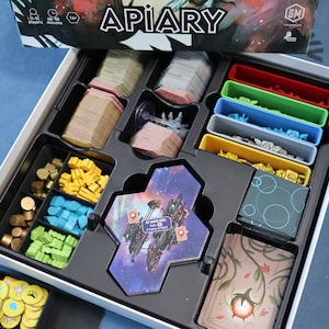 May include: The Apiary board game box is open, revealing a variety of components, including cards, tokens, and tiles. The game is for 1-4 players and takes 60-90 minutes to play. The box is white with a black insert and the game title is printed in black on the box lid.