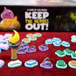 以下が含まれることがあります： 「Keep the Heroes Out!」というゲームで、ハート、サンダル、本、骨、カエルなど、カラフルなゲームピースが使用されています。ゲームピースは赤いフェルトの表面に置かれています。