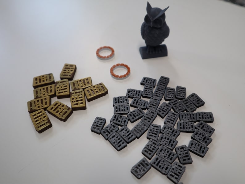 May include: A collection of small, rectangular tiles with a grid pattern. The tiles are in two colors: gold and gray. There is also a small, gray 3D printed owl figurine.