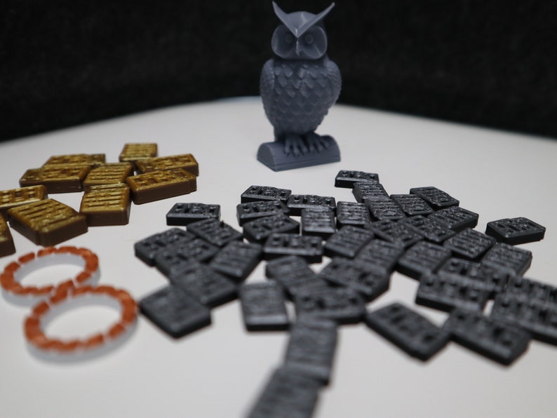 May include: A gray 3D printed owl figurine, a set of brown wooden tiles, and a set of gray plastic tiles. The tiles are scattered on a white surface.