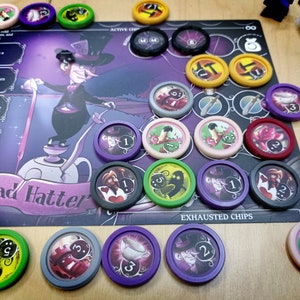 Puede incluir: Un tablero de juego con fondo morado y negro que presenta el texto "Mad Hatter". El tablero tiene varias fichas de juego de colores diferentes con números e imágenes diferentes. Las fichas de juego son moradas, verdes, rojas, negras, grises y amarillas. Las fichas de juego están dispuestas en un círculo alrededor del tablero. El texto "EXHAUSTED CHIPS" está impreso en el tablero.