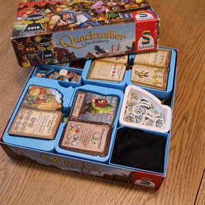 Puede incluir: Caja de juego abierta con el título "Quacksalber von Quedlinburg". La caja, roja y azul, contiene componentes del juego como cartas, fichas y dados, organizados en compartimentos azules. La caja está sobre una superficie de madera.