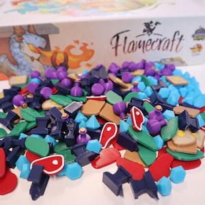 May include: A board game box with the title "Flamecraft" and various game pieces scattered in front. The pieces include blue, red, green, purple, and brown shapes, including leaves, gems, and food items.
