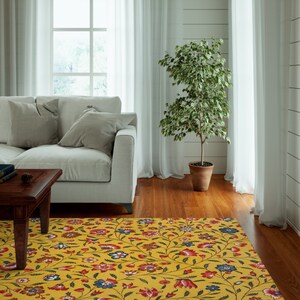 May include: A living room scene featuring a yellow floral rug with red, blue, and white flowers. A white sofa with gray pillows sits near a wooden table. A potted tree stands near a window with white curtains.