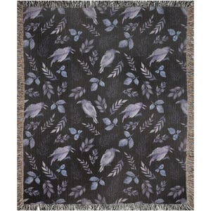 May include: A woven throw blanket featuring a dark charcoal background with a repeating pattern of light gray ravens and various botanical leaf sprigs. The blanket has a textured finish and is finished with a fringed edge on all four sides.