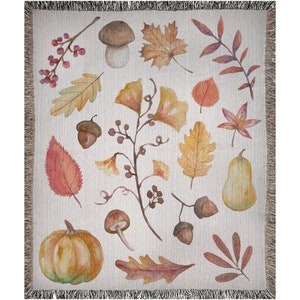 May include: Woven throw blanket featuring a watercolor design of autumn leaves, acorns, mushrooms, and a pumpkin. The blanket has a cream-colored background with a fringed edge. The artwork includes shades of orange, yellow, and brown.