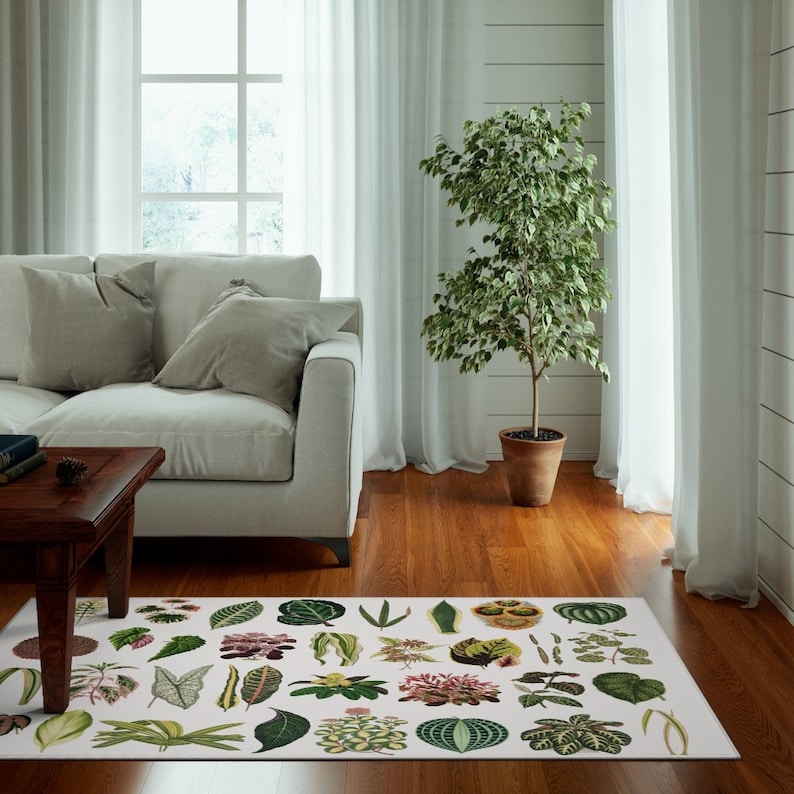 May include: A white area rug with a botanical print featuring various green leaves and flowers. The rug is laid out on a hardwood floor in a living room setting with a white sofa and a potted plant.