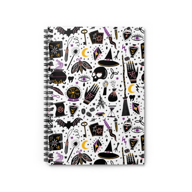 May include: A white spiral notebook with a black and purple pattern of witchy symbols, including a cauldron, a skull, a wand, a broom, a crescent moon, a crystal ball, and a witch's hat.