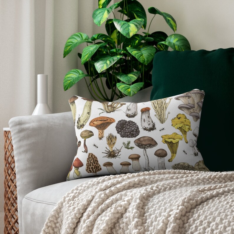 May include: A white throw pillow with a colorful illustration of various mushrooms. The pillow is on a light gray couch with a white throw blanket.
