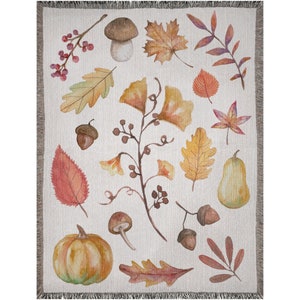 May include: A woven throw blanket featuring a watercolor design of autumn leaves, acorns, mushrooms, and a pumpkin in shades of orange, yellow, and brown. The blanket has a cream-colored background and a fringed edge.