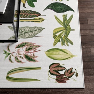 May include: A white rug with a botanical print featuring various green and pink leaves. The leaves are arranged in a repeating pattern and are outlined in black.