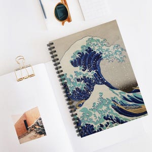 May include: A spiral-bound notebook featuring a print of The Great Wave off Kanagawa on the cover. The notebook is accompanied by a gold-toned metal binder clip, a pair of round-framed sunglasses, and a small square photograph of a building.