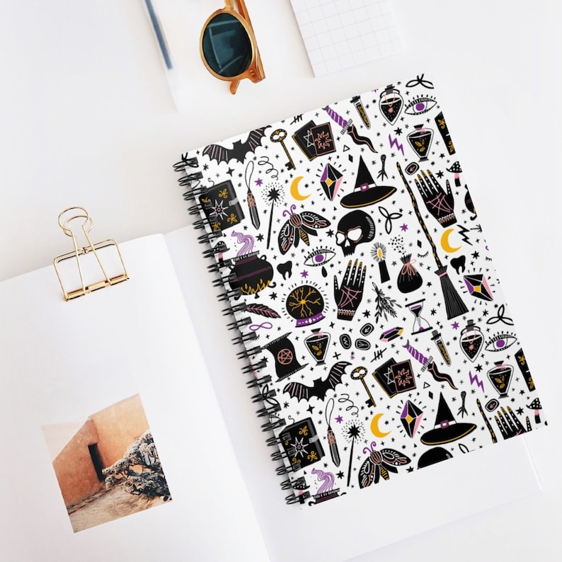May include: A white spiral notebook with a black and white pattern of witchy symbols, including a skull, a cauldron, a wand, a crescent moon, a bat, a crystal, a potion bottle, and a witch's hat. The pattern is repeated throughout the notebook.