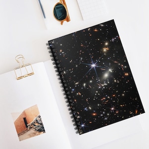 Op de afbeelding: Een zwart spiraalnotitieblok met een galaxy print met een heldere witte ster in het midden, omringd door kleinere sterren en galaxies. Het notitieblok heeft een zwarte spiraalbinding.