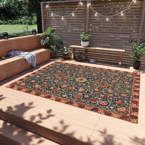May include: A blue and red patterned outdoor rug with floral designs. The rug is laid out on a wooden deck with a bench and plants in the background.