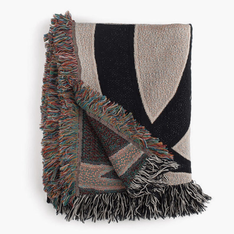 Puede incluir: Manta tejida plegada con un dise&ntilde;o geom&eacute;trico. La manta presenta un patr&oacute;n negro y beige con bordes con flecos multicolores. La manta est&aacute; hecha de un material suave y texturizado, perfecta para agregar calidez y estilo a cualquier espacio habitable.