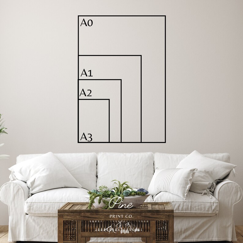 May include: A white wall with four black outlined rectangles labeled A0, A1, A2, and A3. The rectangles are arranged in descending size, with A0 being the largest and A3 being the smallest. The rectangles are positioned on the wall, with A0 at the top and A3 at the bottom. The rectangles are all aligned to the left side of the wall. The wall is a light gray color. A white couch with white pillows is in the foreground. A wooden coffee table with a plant on top is in front of the couch. The coffee table has a sign that says "Pine Print Co."