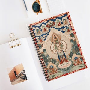 Puede incluir: Un cuaderno de espiral con una ilustración detallada de estilo thangka que muestra una deidad de múltiples brazos rodeada de figuras más pequeñas sobre un fondo azul y crema, con un borde rojo estampado, un clip dorado y gafas de sol.